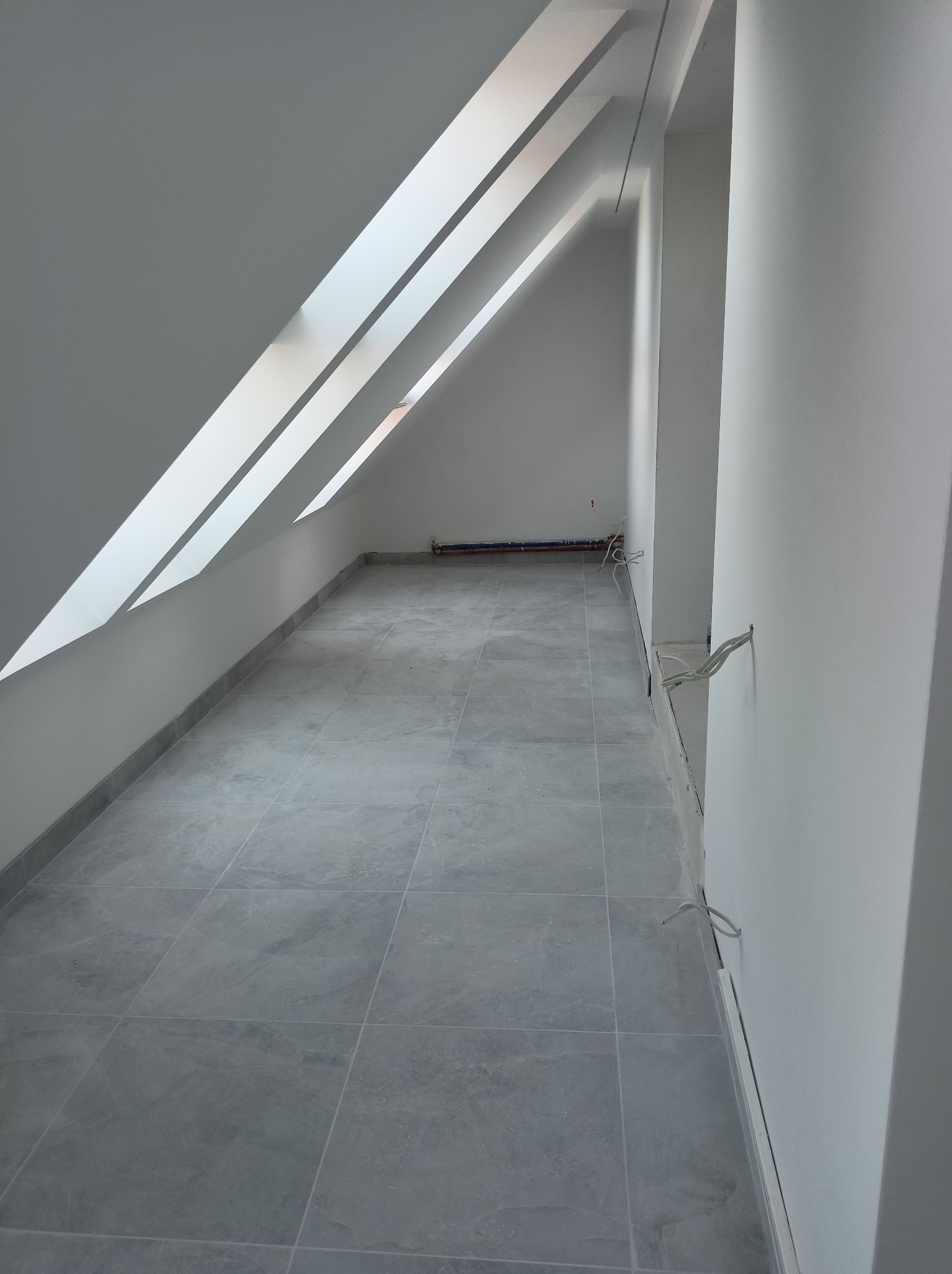 Poddasze z szarymi płytkami podłogowymi, trzema oknami dachowymi w skośnym suficie i wystającymi przewodami elektrycznymi ze ściany.