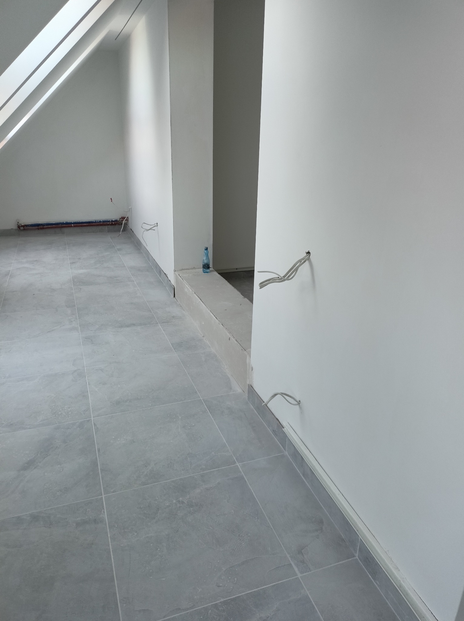 Szare płytki podłogowe w surowym wnętrzu poddasza ze skosami, widoczne kable wystające ze ściany i rury grzewcze przy podłodze, butelka wody na podeście.