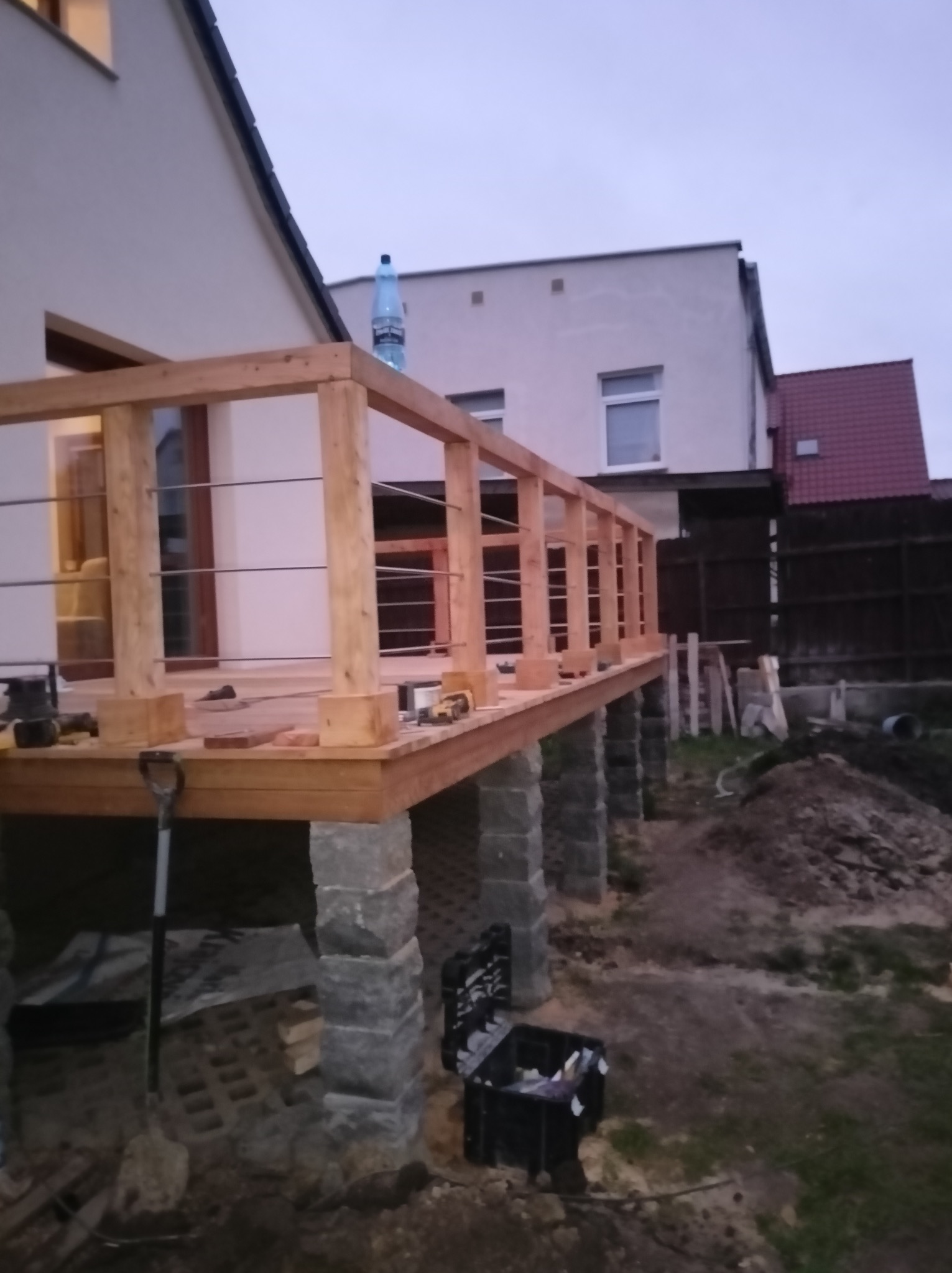 Budowa drewnianego tarasu na kamiennych podporach, widoczna butelka wody na balustradzie, narzędzia i skrzynka narzędziowa na podłodze, dom w tle.