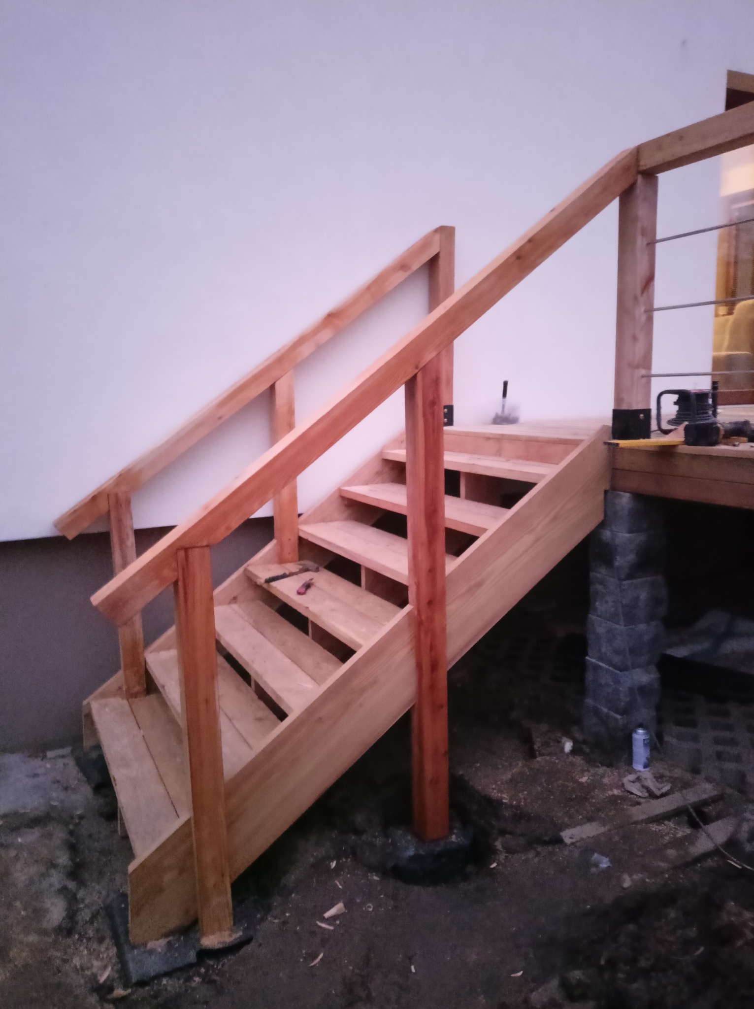 Drewniane schody zewnętrzne w trakcie budowy z widocznymi narzędziami na stopniach i podestem z szarym wykończeniem.