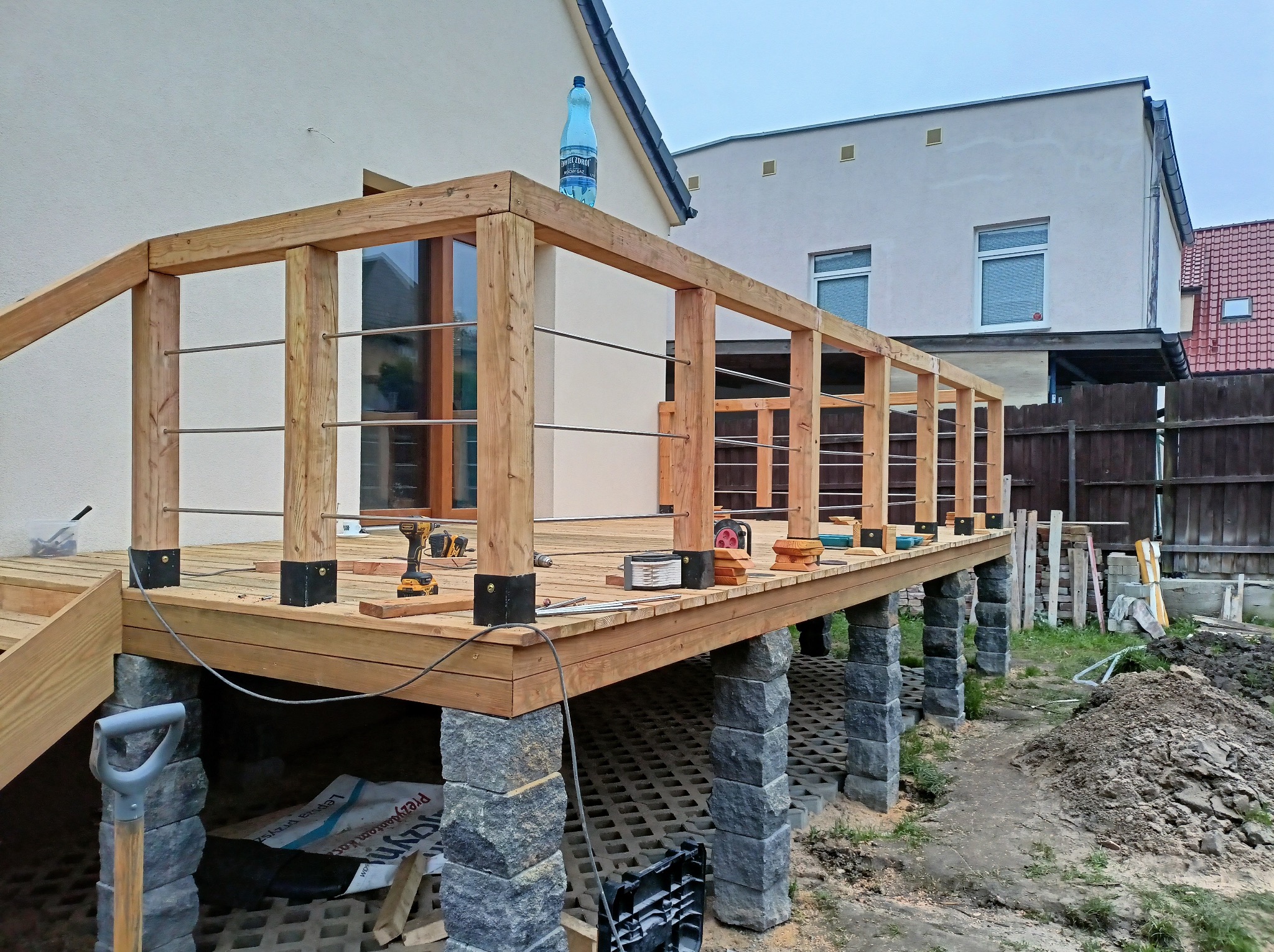 Drewniany taras w trakcie budowy, wsparty na kamiennych filarach, widoczne narzędzia, butelka wody na górnej krawędzi konstrukcji.