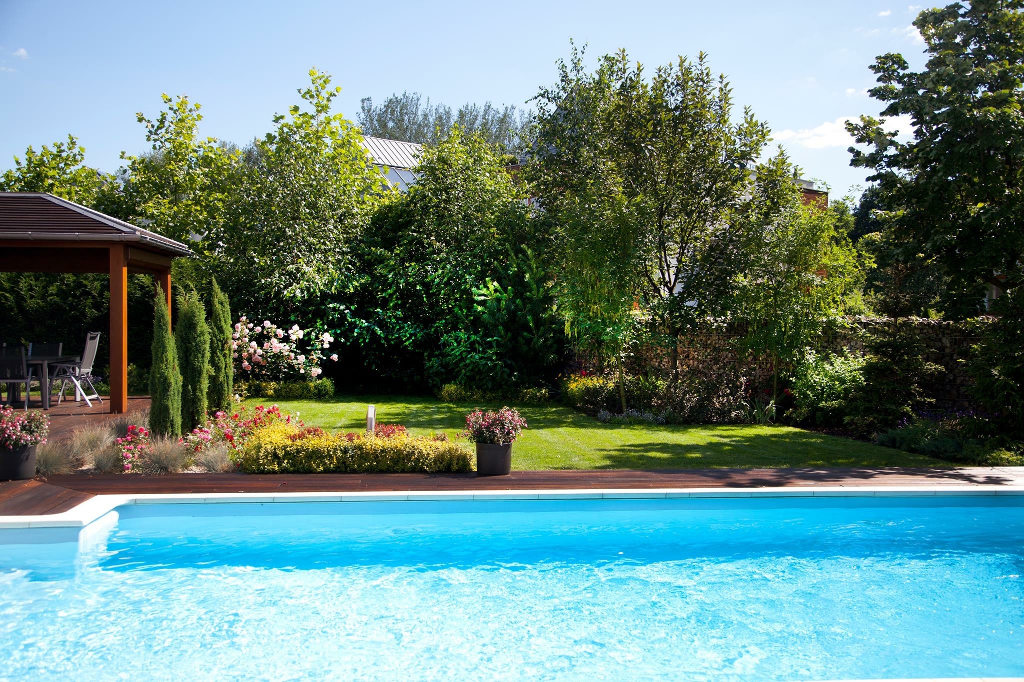 Błękitny basen z drewnianym tarasem, otoczony bujną zielenią ogrodu z altaną i kwitnącymi rabatami w słoneczny dzień.