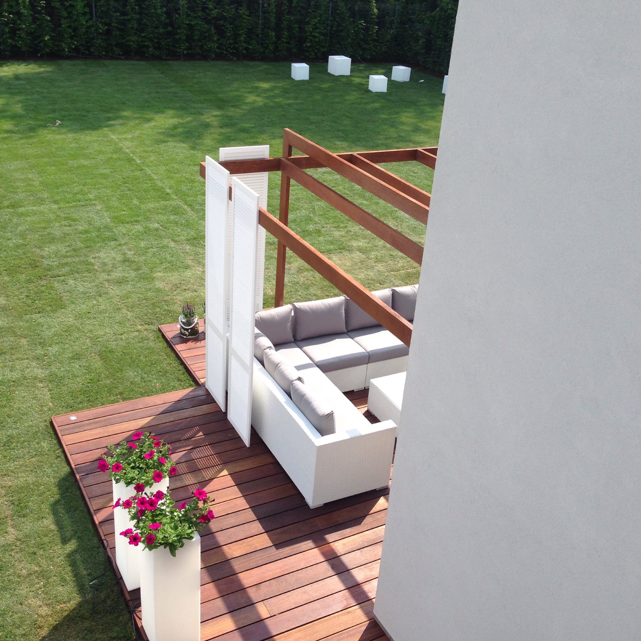 Nowoczesny taras z drewnianą podłogą, białymi meblami ogrodowymi i pergolą, widok z góry na zadbany trawnik z dekoracyjnymi białymi kostkami.