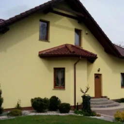 Żółta elewacja domu jednorodzinnego z brązowym dachem i drewnianymi elementami wykończeniowymi, widok z poziomu gruntu na zadbany ogród. Widoczne schody wejściowe z szarego kamienia prowadzące...