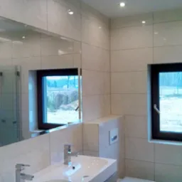 Wykończona łazienka z beżowymi płytkami, lustrem odbijającym kabinę prysznicową i oknem z widokiem na zewnątrz.