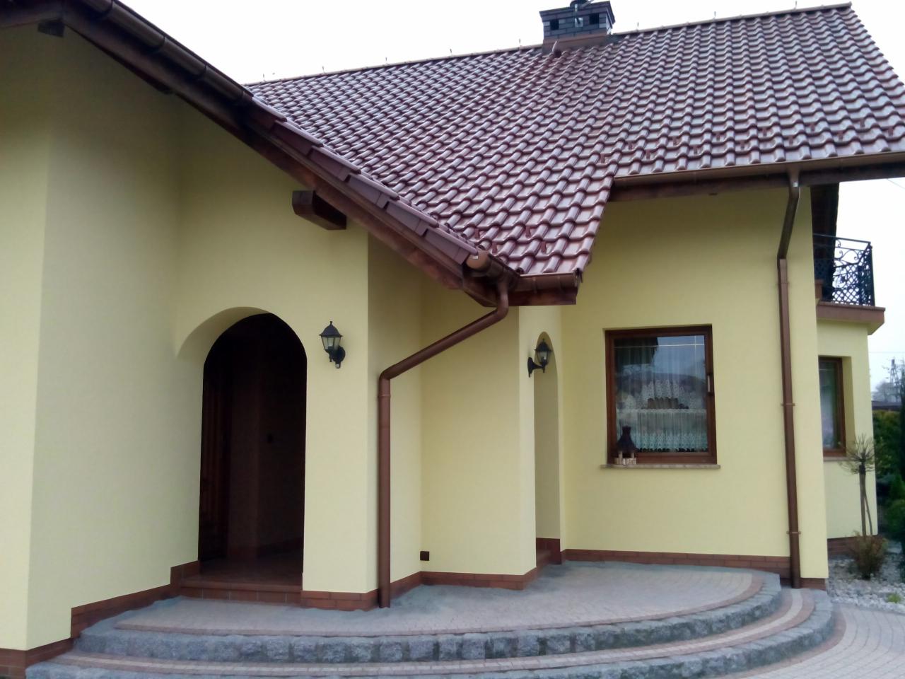 Żółta elewacja domu jednorodzinnego z brązową dachówką, widoczne oświetlenie zewnętrzne, rynny i kamienne schody wejściowe.