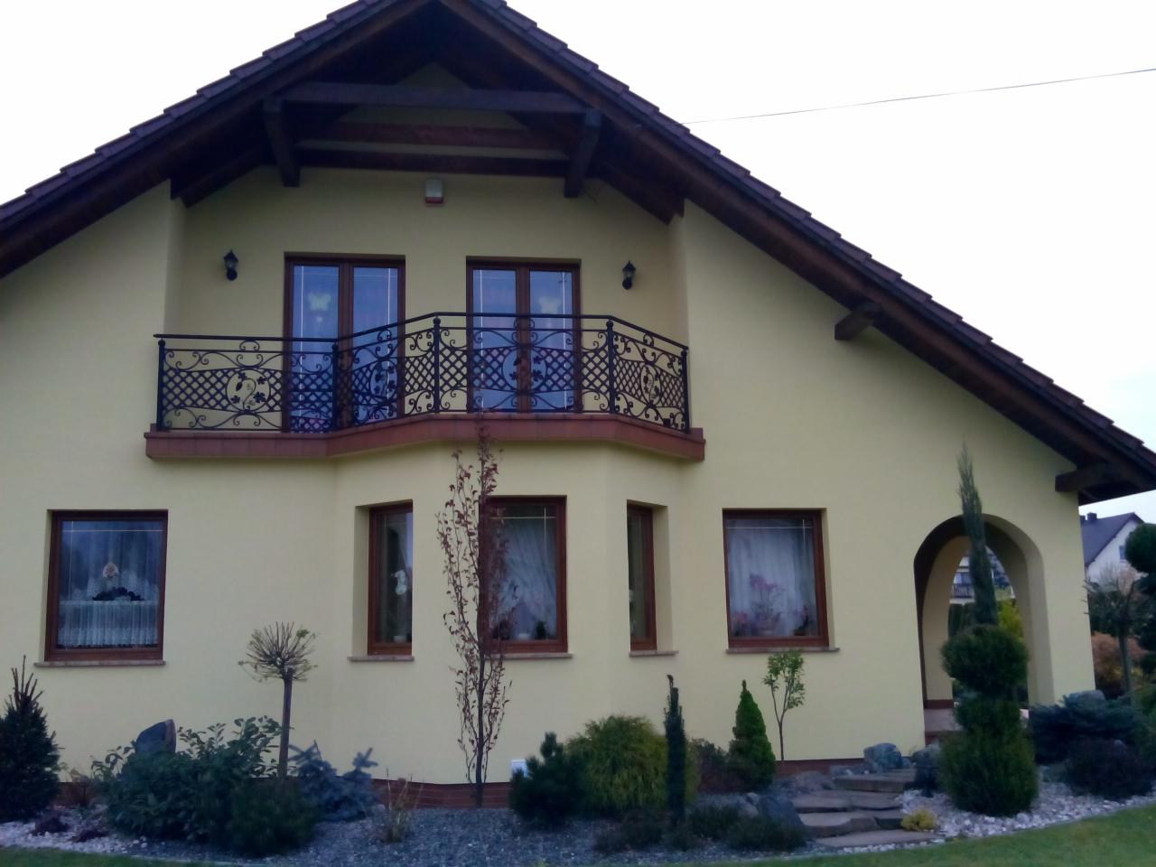 Dwupiętrowy dom z żółtą elewacją, balkonem z ozdobną, metalową balustradą i łukowym wejściem, otoczony zadbanym ogrodem z krzewami i drzewami iglastymi.
