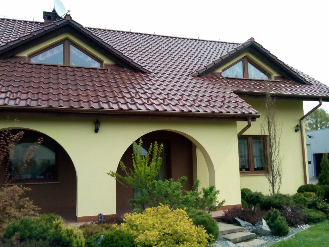 Jasnożółty dom jednorodzinny z brązowym dachem z dachówki, łukowym wejściem i zadbanym ogrodem z roślinami o różnych kolorach i fakturach, widoczne okna trójkątne na poddaszu.