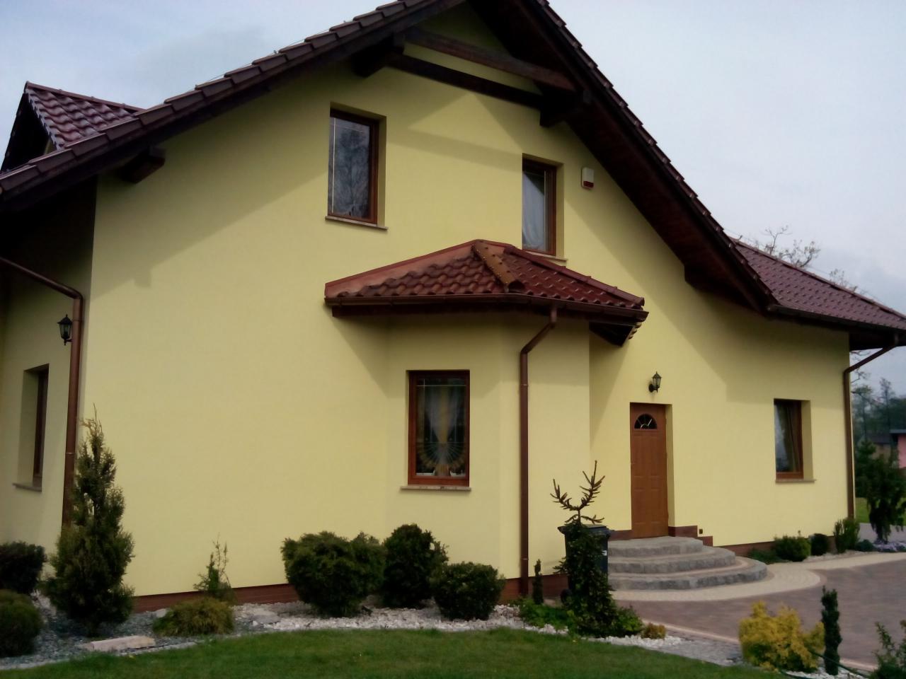 Żółta elewacja domu jednorodzinnego z brązowym dachem i drewnianymi elementami wykończeniowymi, widok z poziomu gruntu na zadbany ogród. Widoczne schody wejściowe z szarego kamienia prowadzące...