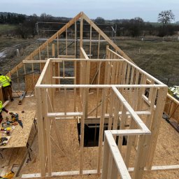 MonterBud - Konstrukcja szkieletowa domu w trakcie budowy, widok z góry na drewniany szkielet, dwóch pracowników w odblaskowych kurtkach przy stole z narzędziami, krajobraz wiejski w tle.