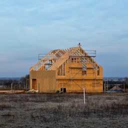 MonterBud - Wznoszona konstrukcja domu z drewna i płyt OSB na tle porannego nieba, widoczne rusztowania i elementy konstrukcyjne dachu, w tle farma wiatrowa.