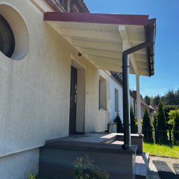 MonterBud - Wejście do domu z zadaszeniem i drewnianym podestem, widok z boku w słoneczny dzień. Zauważalny okrągły otwór okienny i czarna rynna.