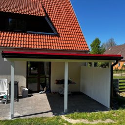 MonterBud - Zadaszenie tarasu z białymi słupami i ścianką działową z pionowych desek, widok na dom z czerwoną dachówką i zielony ogród w słoneczny dzień.