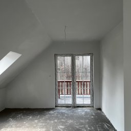 MonterBud - Poddasze po malowaniu na biało, widoczne okno balkonowe z drewnianą balustradą balkonu, surowa podłoga przed wykończeniem, wystający kabel oświetleniowy.