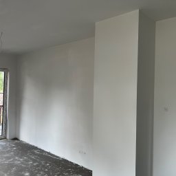 MonterBud - Wnętrze w trakcie remontu: świeżo pomalowane na biało ściany, z widocznymi śladami po remoncie na podłodze, okno balkonowe z widokiem na zewnątrz i surowa ościeżnica drzwiowa.