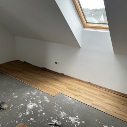 MonterBud - Początek układania paneli podłogowych w pokoju na poddaszu ze skośnym sufitem i oknem dachowym. Na szarym, surowym podłożu widoczne białe ślady po zaprawie. Część paneli już ułożona, przy ścianie...