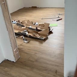 MonterBud - Poddasze w trakcie wykańczania, z częściowo ułożoną podłogą z paneli w odcieniach brązu, narzędzia i resztki opakowań po panelach na podłodze, okno dachowe w tle.