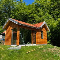 MonterBud - Nowoczesny domek z drewnianą elewacją i czerwoną dachówką, otoczony zielenią, z widocznymi śladami prac wykończeniowych na trawniku.
