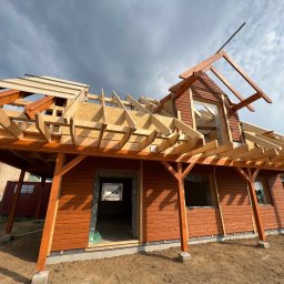 MonterBud - Konstrukcja drewnianego domu w trakcie budowy, widoczne elementy szkieletu dachu i ścian, z brązową elewacją, na tle pochmurnego nieba.