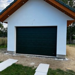 MonterBud - Nowo wybudowany garaż z ciemnozieloną bramą segmentową, wykończony białą elewacją i drewnianą podbitką dachu, widoczne płyty gipsowe na trawie.