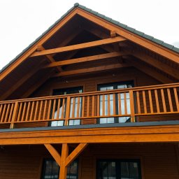 MonterBud - Dwupiętrowy dom z drewna z balkonem, pomalowany na ciepły, miodowy odcień brązu, widok z dołu ukazujący konstrukcję dachu i czarne ramy okienne.