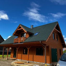 MonterBud - Nowo wybudowany, dwukondygnacyjny dom z bali w odcieniach brązu z ciemnozielonym dachem na tle błękitnego nieba z białymi obłokami. Widoczny balkon z drewnianą balustradą, rynna i fragment...