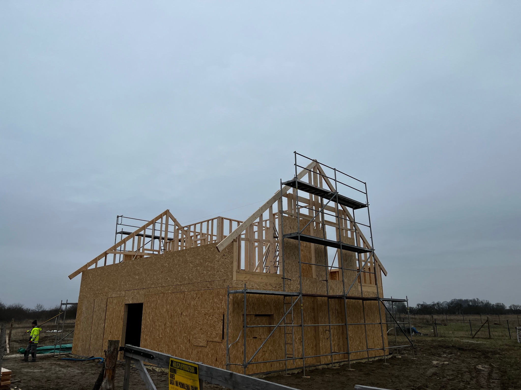 Konstrukcja domu w stanie surowym otwartym z płyt OSB, z widocznym szkieletem dachu i rusztowaniami, pracownik w odblaskowej kamizelce w tle.