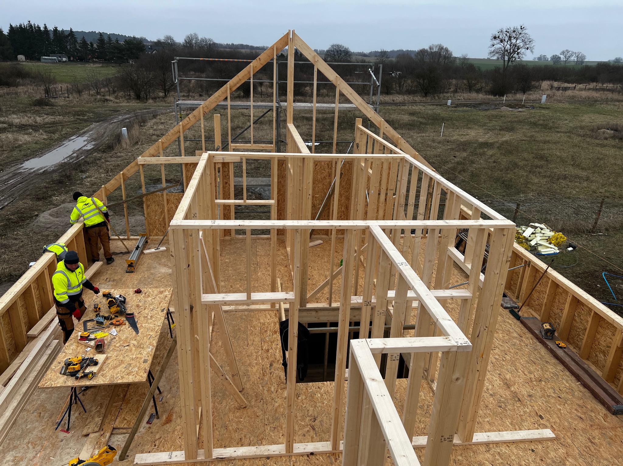Konstrukcja szkieletowa domu w trakcie budowy, widok z góry na drewniany szkielet, dwóch pracowników w odblaskowych kurtkach przy stole z narzędziami, krajobraz wiejski w tle.