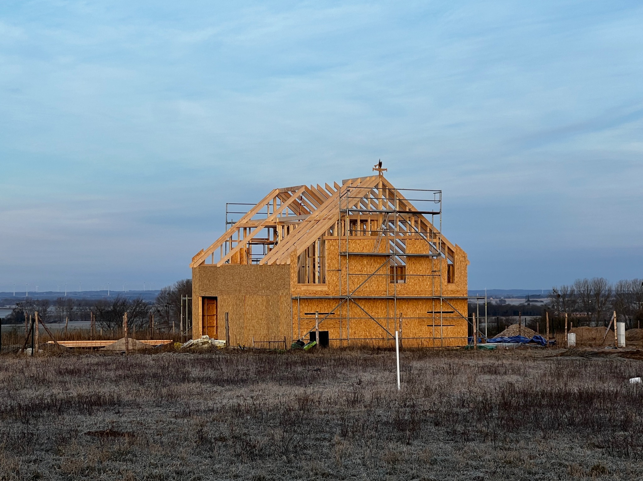 Wznoszona konstrukcja domu z drewna i płyt OSB na tle porannego nieba, widoczne rusztowania i elementy konstrukcyjne dachu, w tle farma wiatrowa.