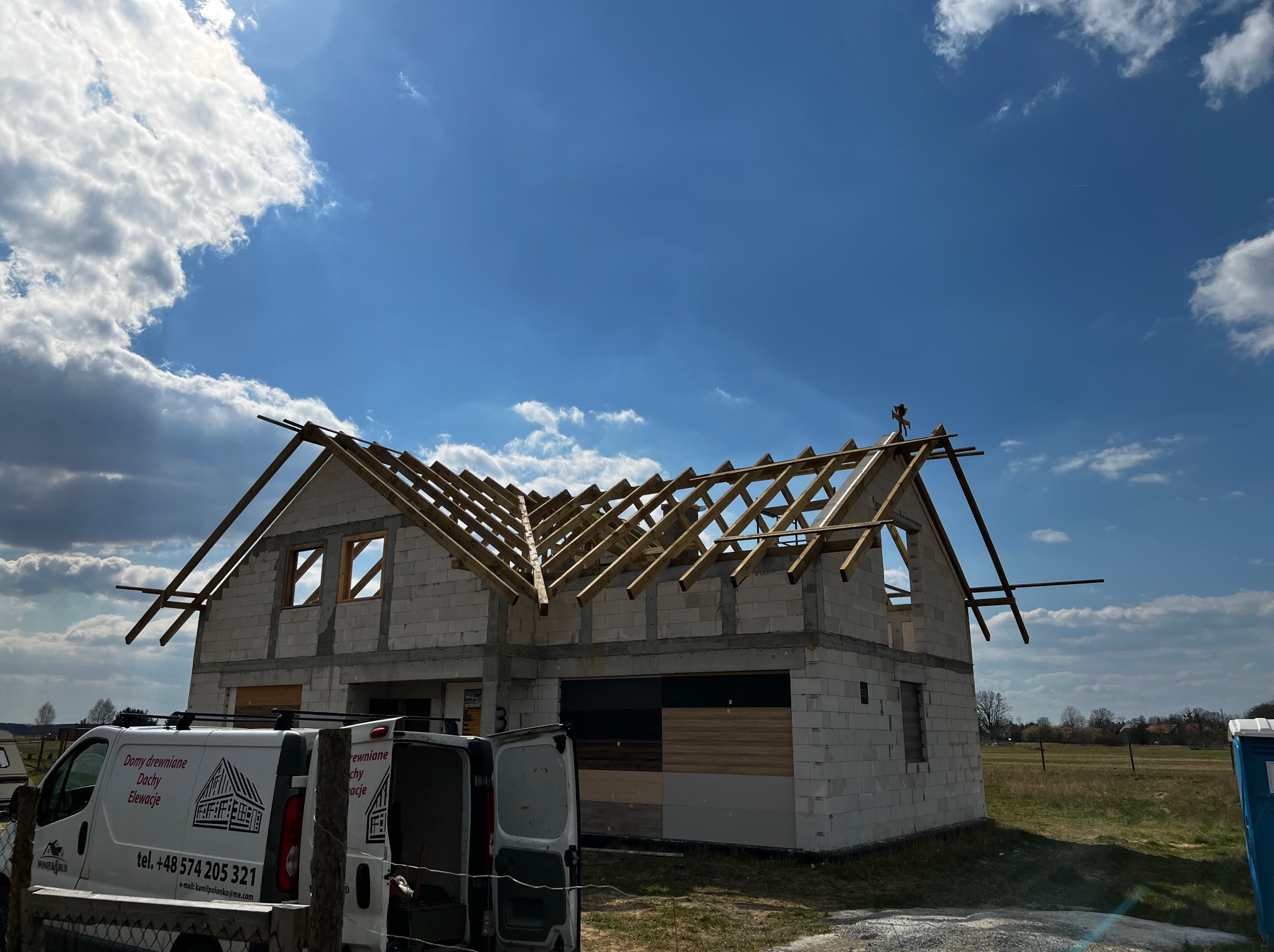 Niedokończony budynek z białych bloczków z drewnianą konstrukcją dachu w słoneczny dzień, samochód dostawczy firmy budowlanej z widocznym numerem telefonu w tle.