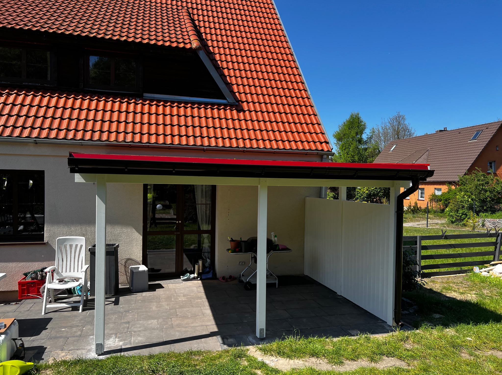 Zadaszenie tarasu z białymi słupami i ścianką działową z pionowych desek, widok na dom z czerwoną dachówką i zielony ogród w słoneczny dzień.