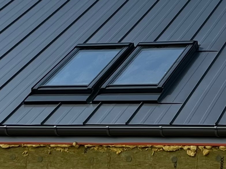Dwa okna dachowe w ciemnoszarym, metalowym dachu skośnym. Widoczna izolacja pod okapem.