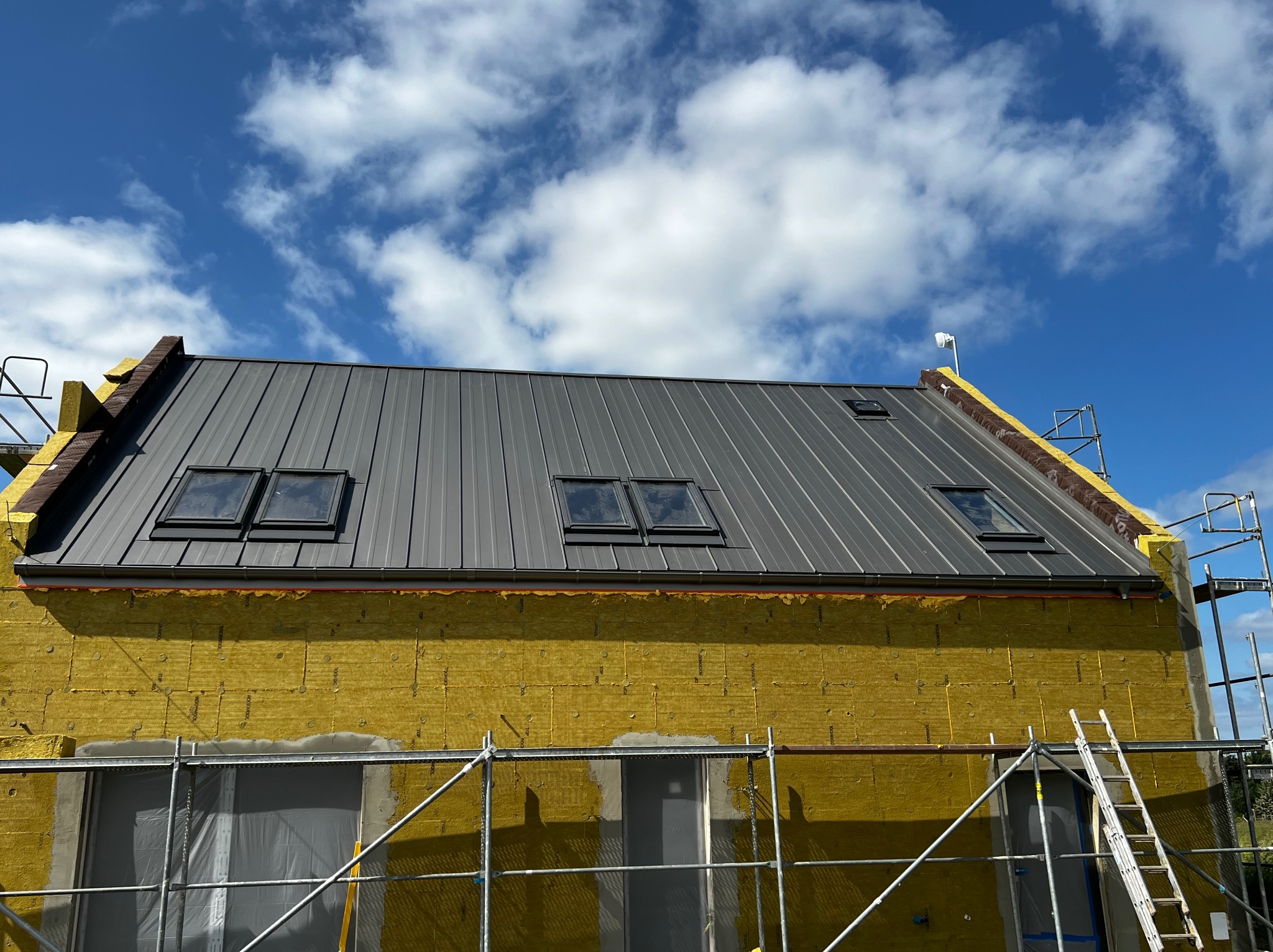 Dom w trakcie ocieplania, widoczna żółta wełna mineralna na ścianach, szare metalowe pokrycie dachu z oknami dachowymi, rusztowania i drabina w tle błękitne niebo z chmurami.