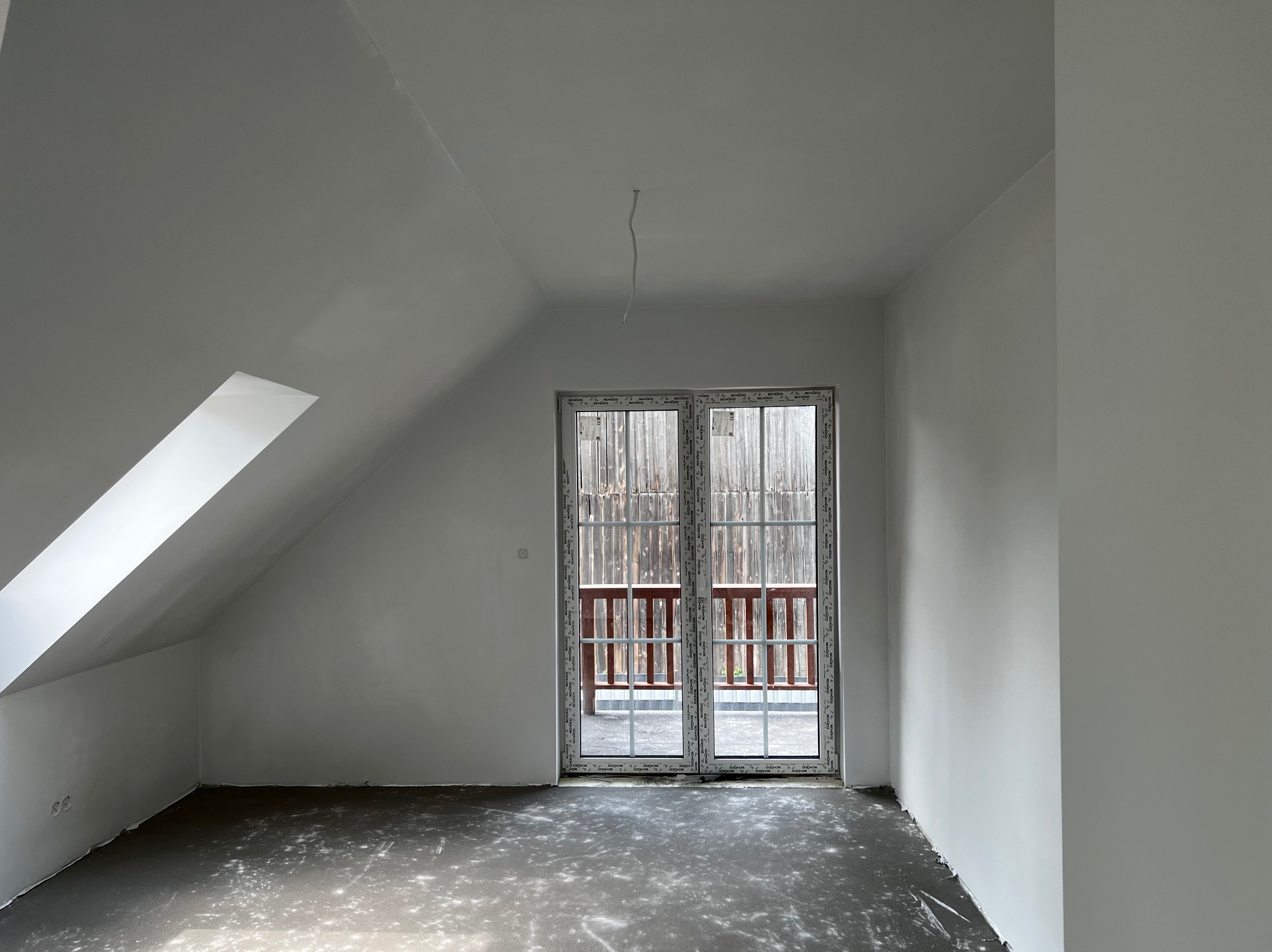 Poddasze po malowaniu na biało, widoczne okno balkonowe z drewnianą balustradą balkonu, surowa podłoga przed wykończeniem, wystający kabel oświetleniowy.