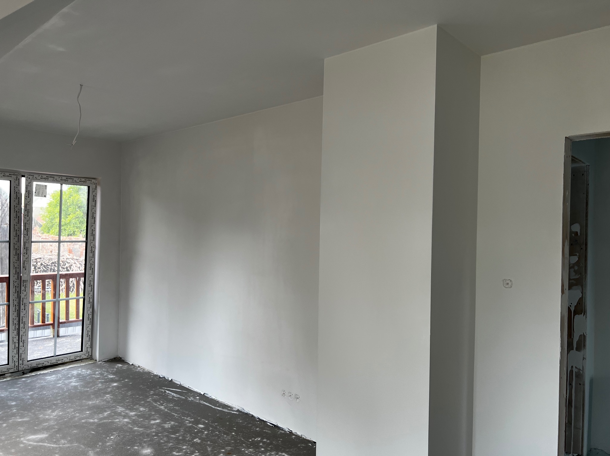 Wnętrze w trakcie remontu: świeżo pomalowane na biało ściany, z widocznymi śladami po remoncie na podłodze, okno balkonowe z widokiem na zewnątrz i surowa ościeżnica drzwiowa.