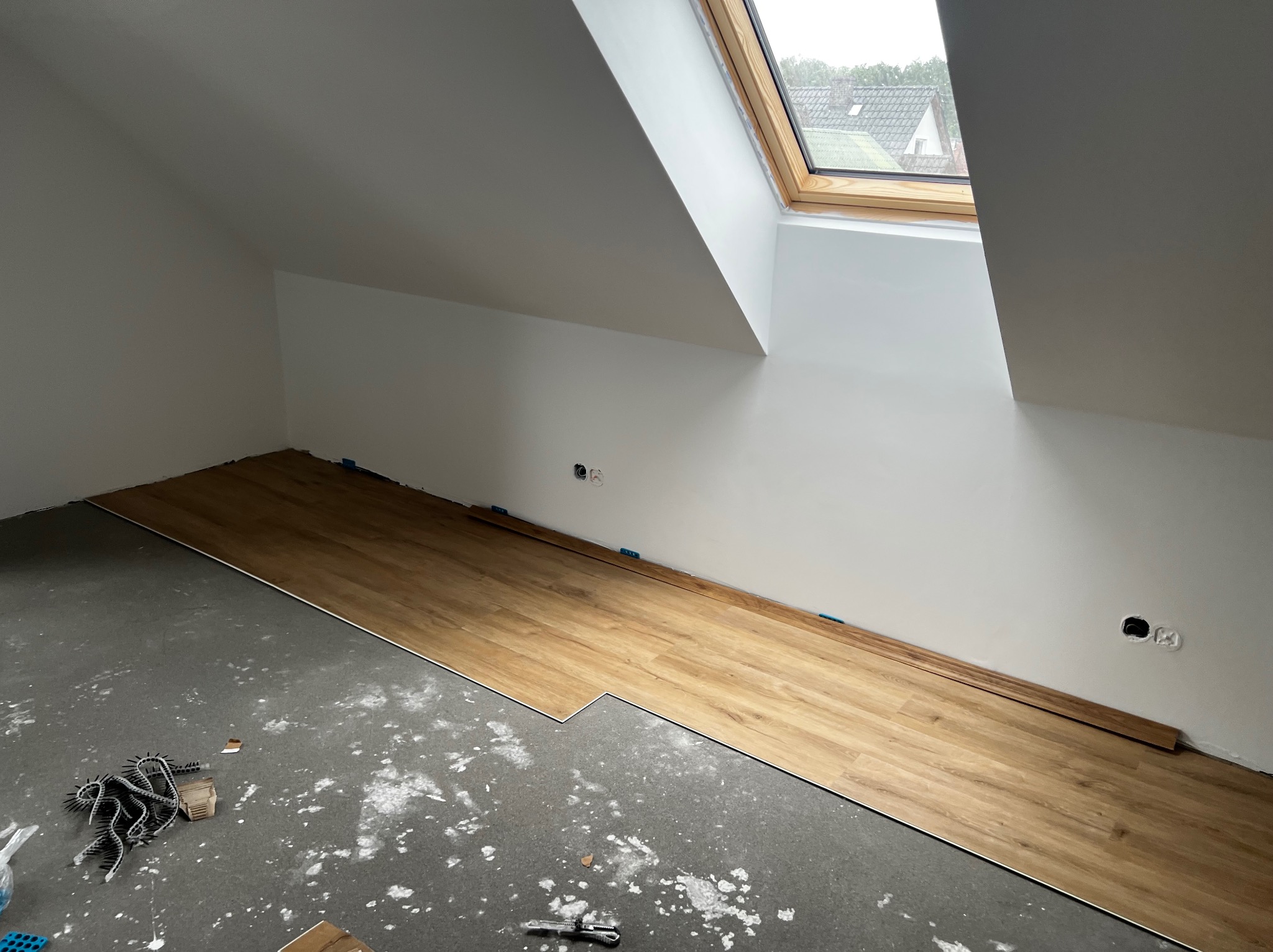 Początek układania paneli podłogowych w pokoju na poddaszu ze skośnym sufitem i oknem dachowym. Na szarym, surowym podłożu widoczne białe ślady po zaprawie. Część paneli już ułożona, przy ścianie...