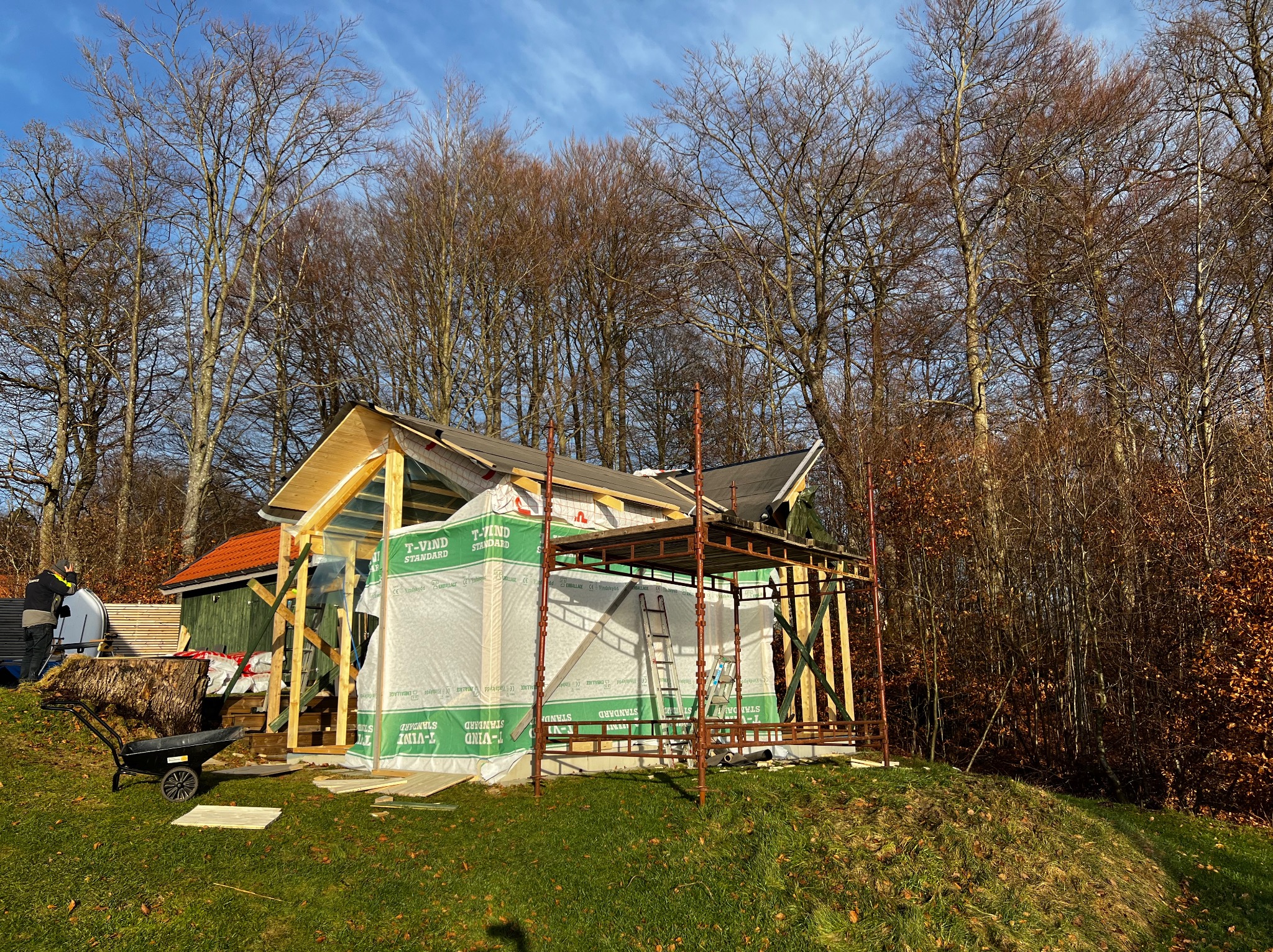 Budowa szkieletowej konstrukcji domu w Stepnicy, widoczne rusztowania, folia ochronna T-Vind Standard, częściowo wykończony dach i przygotowane deski, w tle drzewa.