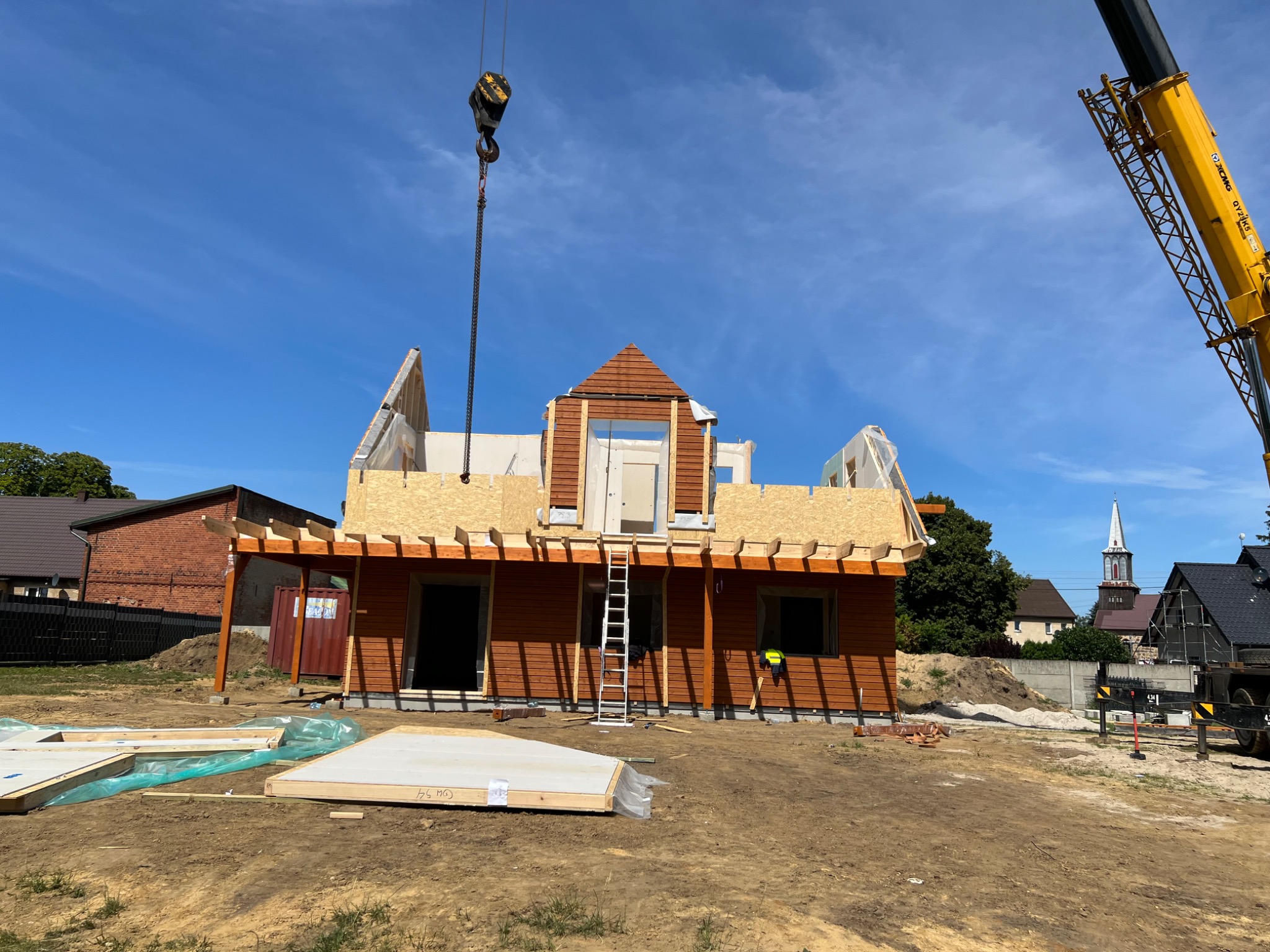 Budowa szkieletowego domu drewnianego z użyciem dźwigu; widoczne elementy konstrukcyjne, ściany z płyt OSB, drewniane belki, drabina oparta o ścianę, a w tle kościół z wieżą.