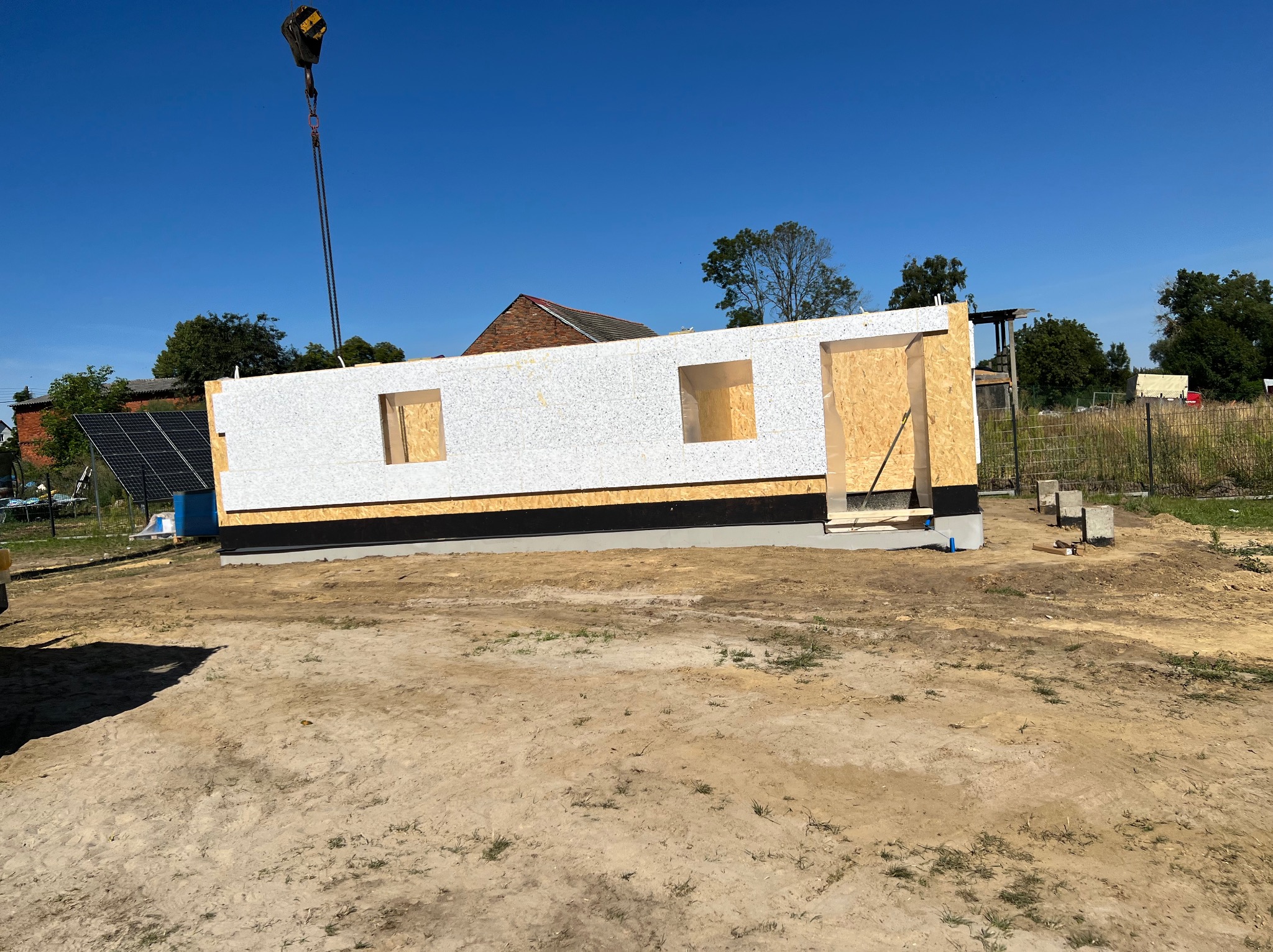 Wznoszenie szkieletu domu z płyty warstwowej, widoczny fragment ściany z otworami okiennymi, podnoszony dźwigiem na tle błękitnego nieba i zabudowań gospodarczych.