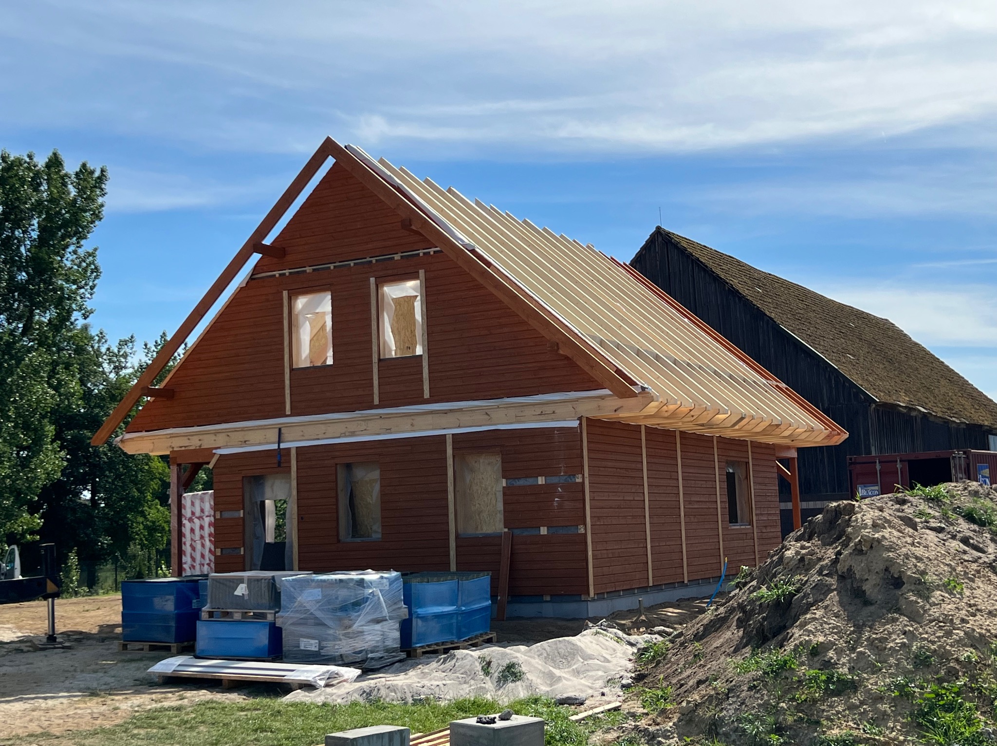 Budowa domu z drewnianą elewacją, widoczne elementy konstrukcji dachu, okna zabezpieczone folią, niebieskie beczki i materiały budowlane przed budynkiem, w tle stary budynek gospodarczy.