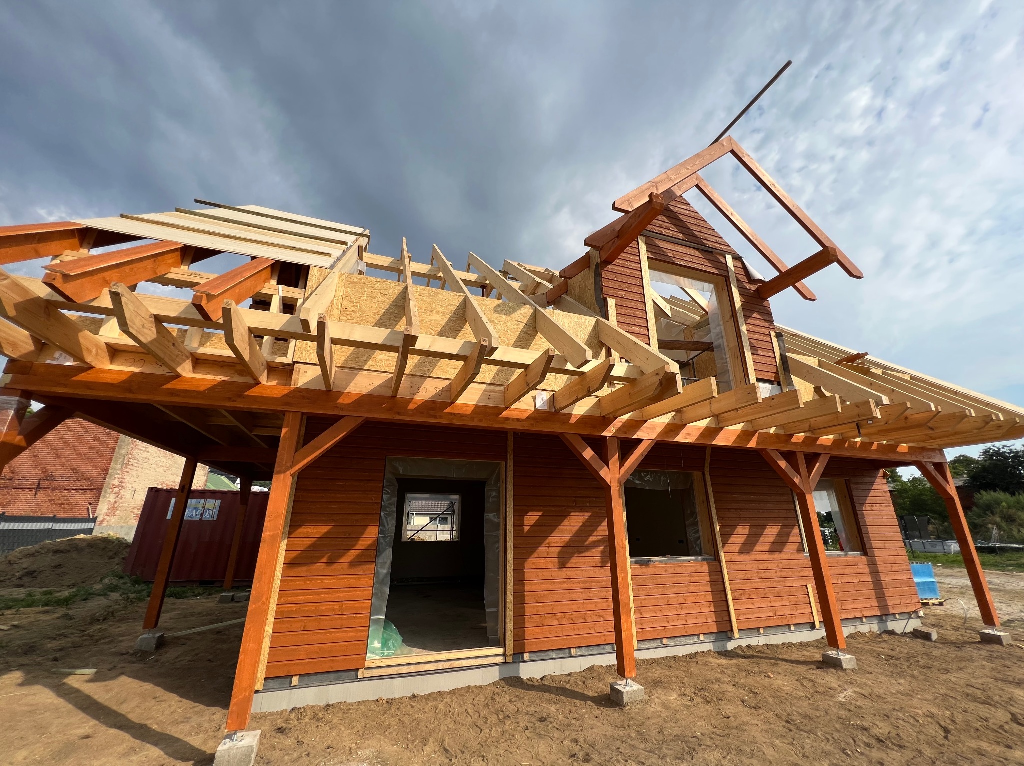 Konstrukcja drewnianego domu w trakcie budowy, widoczne elementy szkieletu dachu i ścian, z brązową elewacją, na tle pochmurnego nieba.