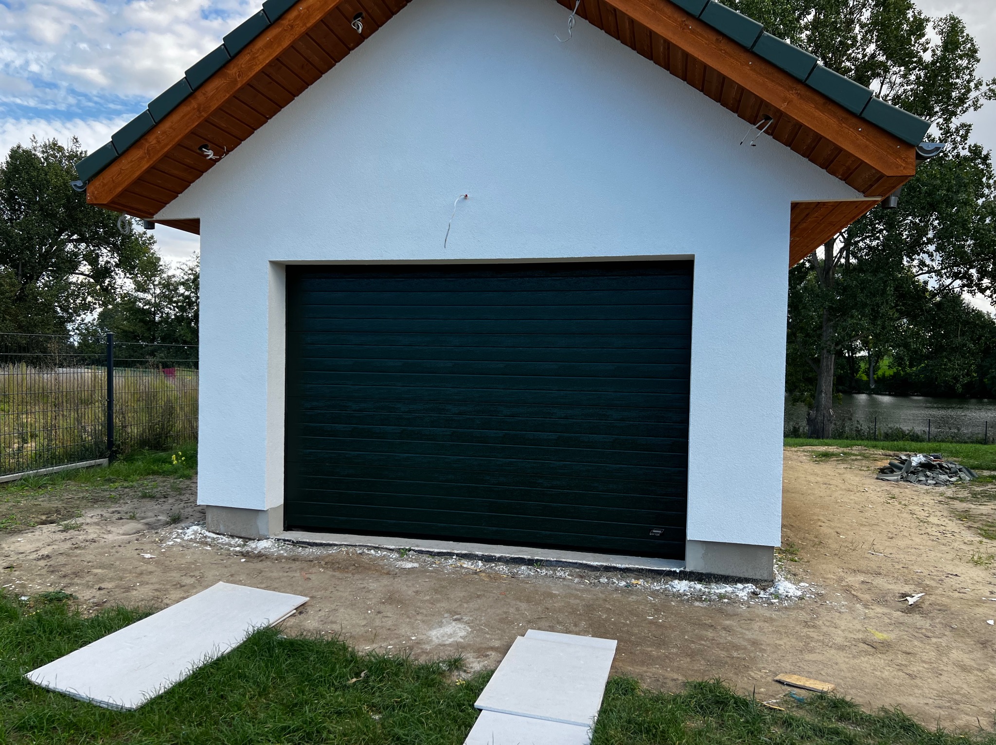 Nowo wybudowany garaż z ciemnozieloną bramą segmentową, wykończony białą elewacją i drewnianą podbitką dachu, widoczne płyty gipsowe na trawie.