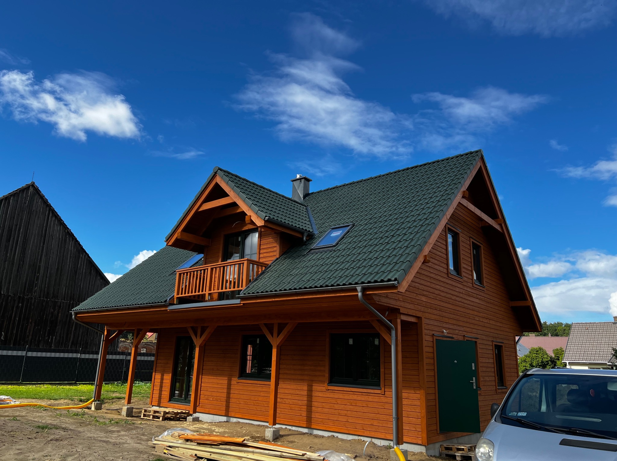 Nowo wybudowany, dwukondygnacyjny dom z bali w odcieniach brązu z ciemnozielonym dachem na tle błękitnego nieba z białymi obłokami. Widoczny balkon z drewnianą balustradą, rynna i fragment...