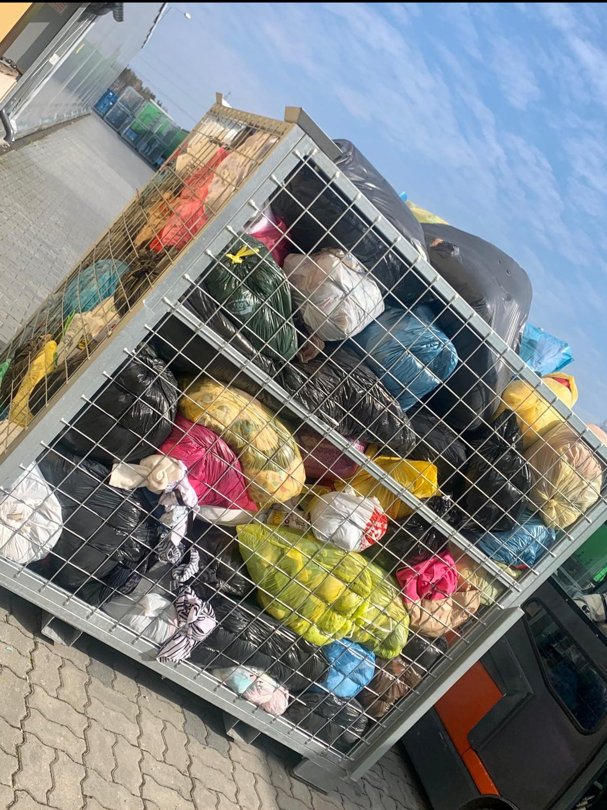 Metalowy kontener siatkowy wypełniony workami z odzieżą, widoczne różne kolory i faktury materiałów, tło stanowi kostka brukowa i fragment nieba.