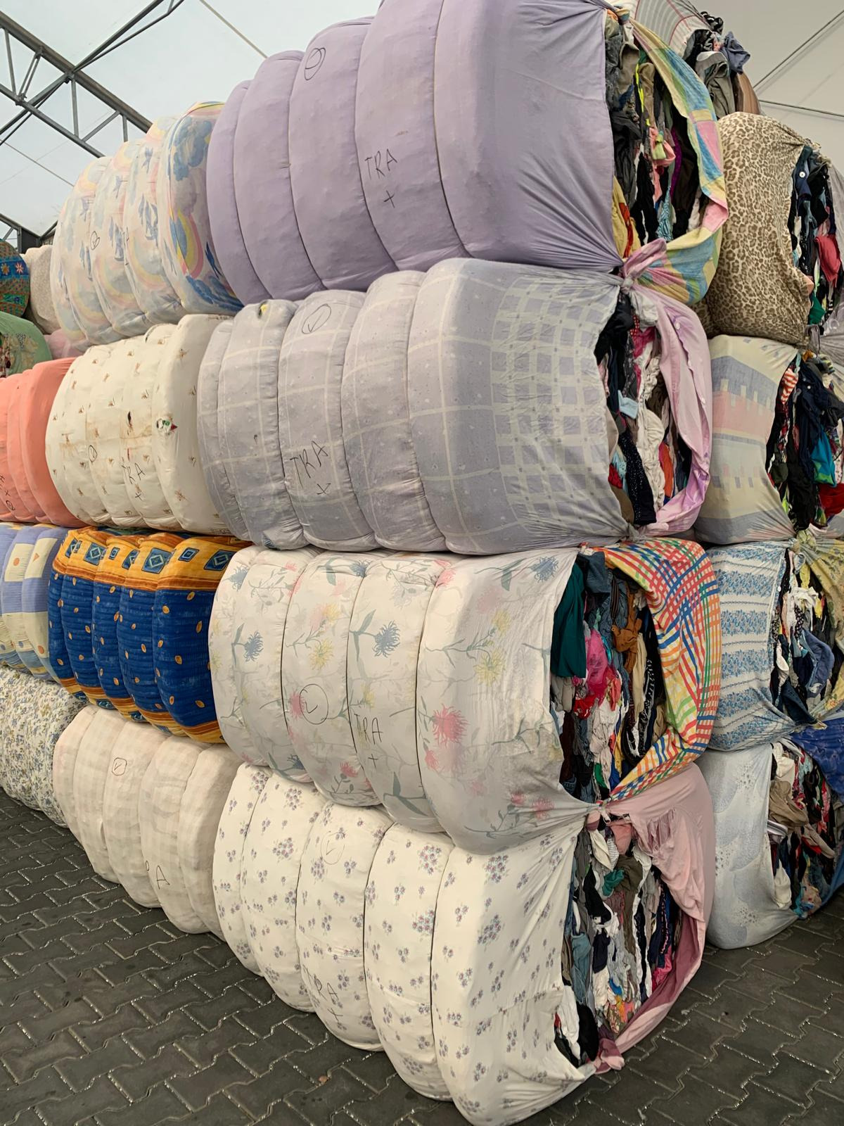 Stos sprasowanych bel z odzieżą używaną w workach z różnymi wzorami, część worków z widoczną zawartością, niektóre z napisem 'TRA Bales'.