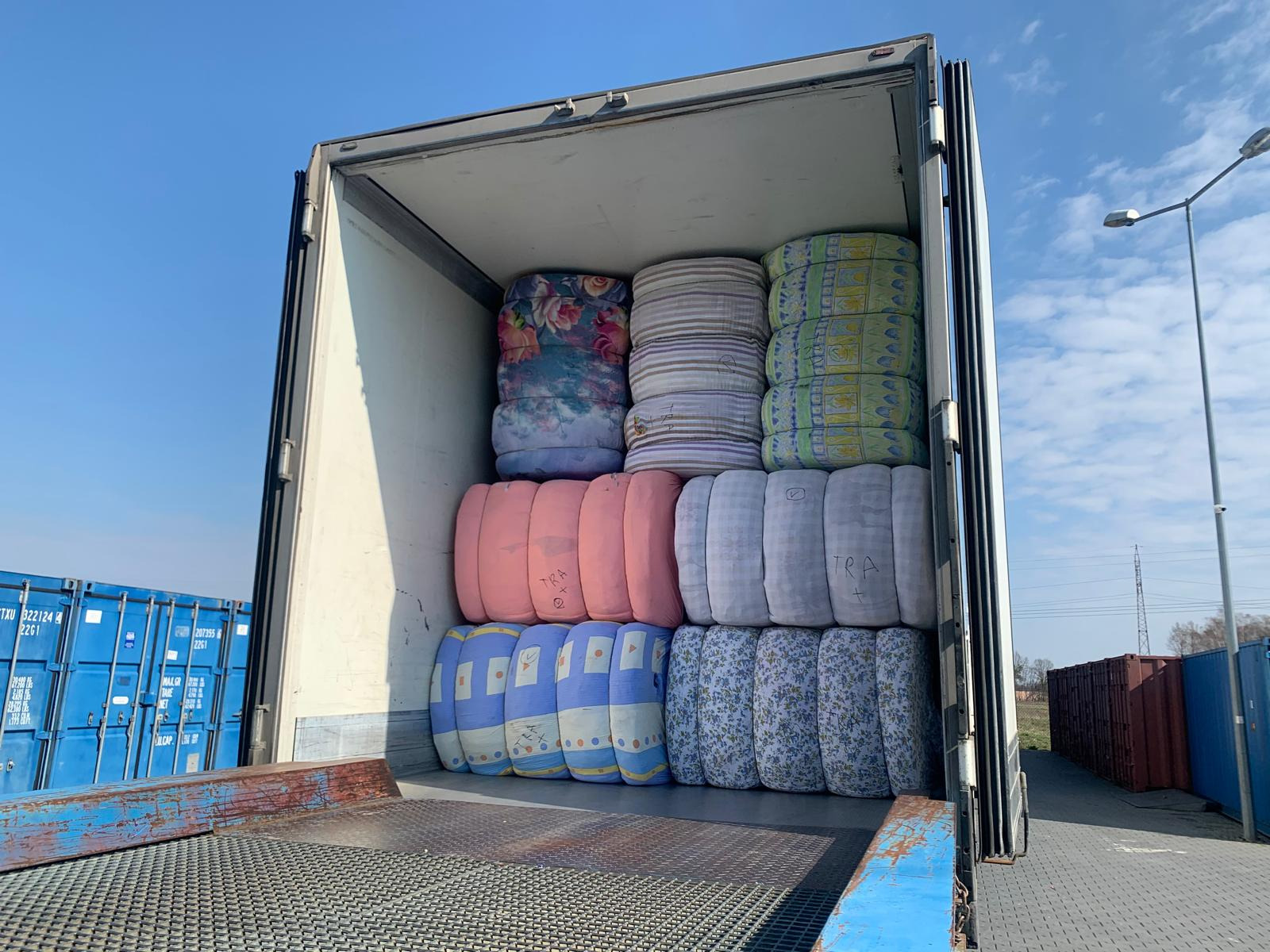Wnętrze naczepy wypełnionej sprasowanymi belami tekstyliów w różnych wzorach, widoczne niebieskie kontenery w tle.