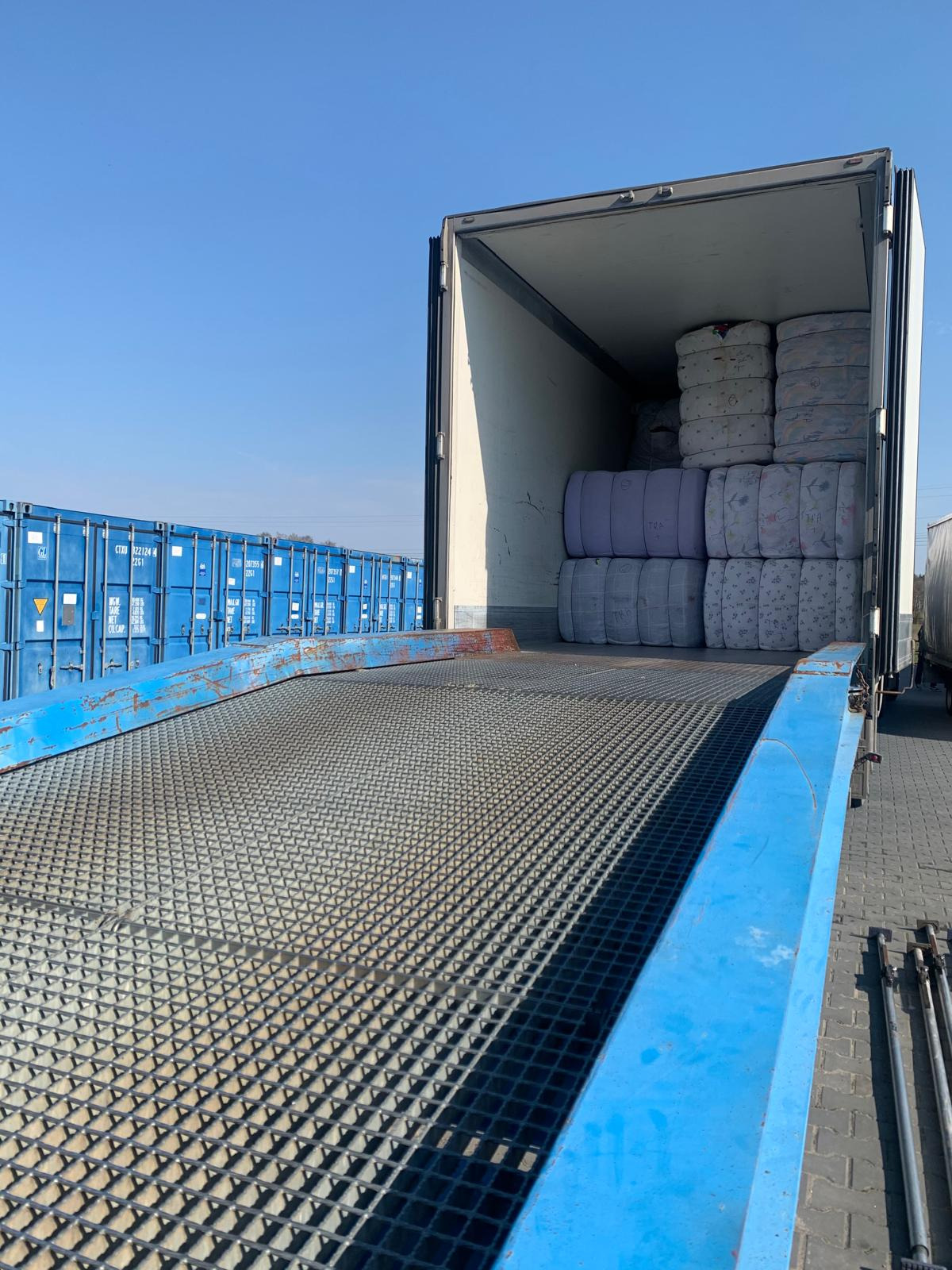 Wnętrze kontenera wypełnione belami tekstyliów w różnych odcieniach, widok z rampy załadunkowej z metalową kratą, w tle niebieskie kontenery transportowe.