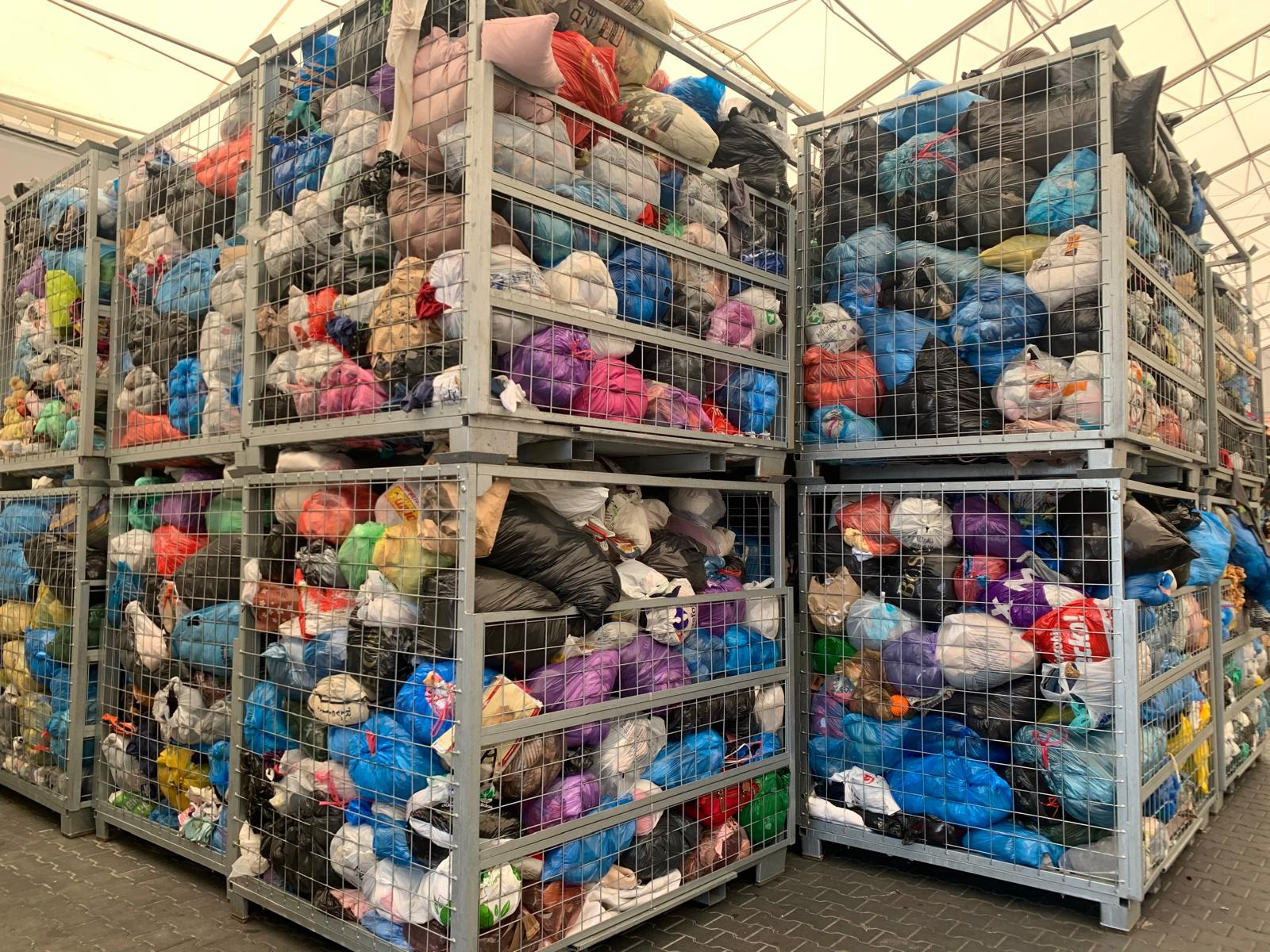 Magazyn odzieży używanej: metalowe kontenery wypełnione workami z odzieżą, widoczne kolory niebieski, fioletowy, czarny i biały, oświetlenie magazynowe.