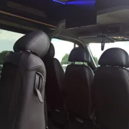 Wnętrze luksusowego busa do przewozu osób, widoczne czarne skórzane fotele z zagłówkami, oświetlenie LED na suficie i zamontowany telewizor.