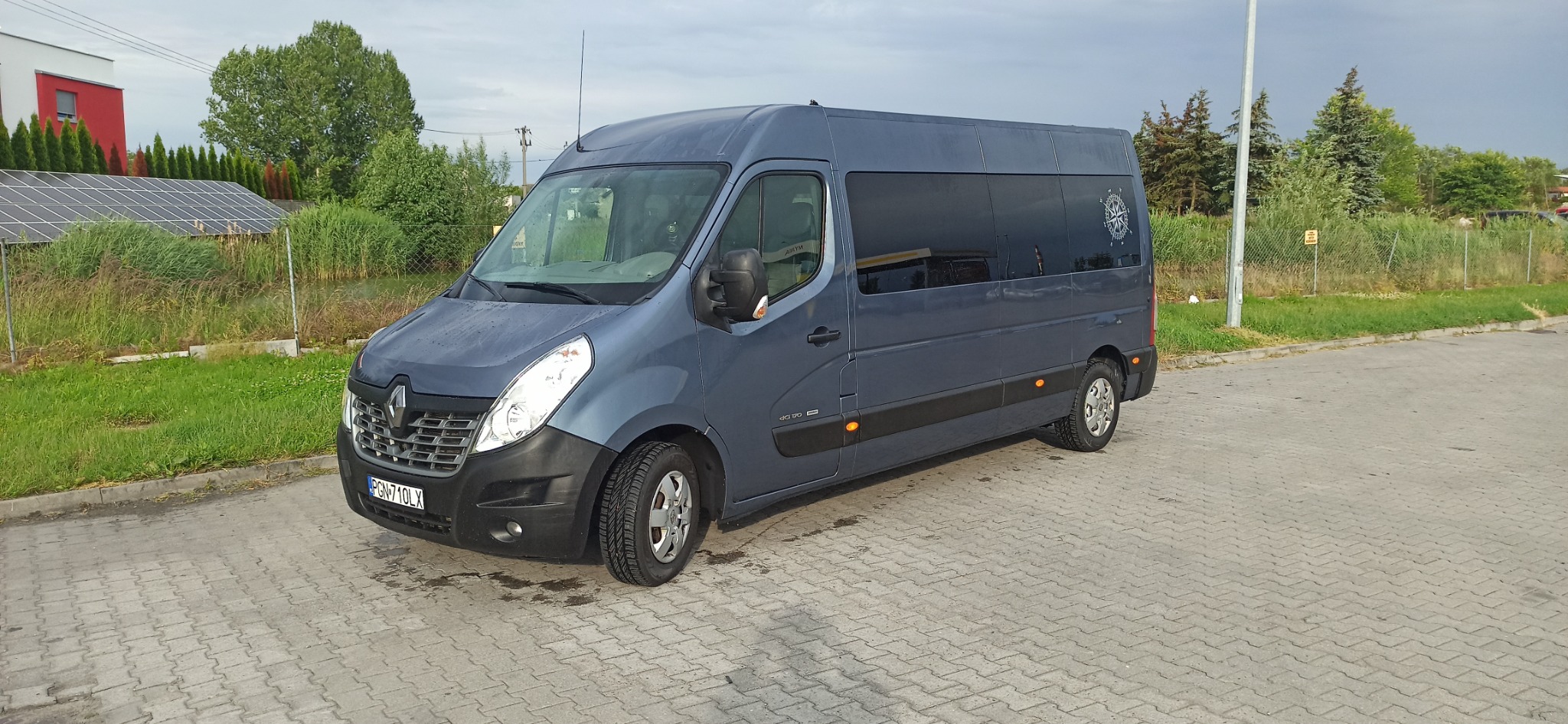 Szary Renault Master z przyciemnianymi szybami i naklejoną różą wiatrów na tylnej szybie, zaparkowany na kostce brukowej.